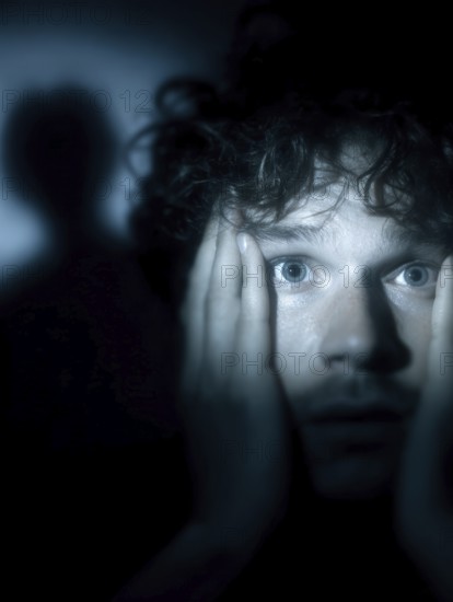 An anxious man with wide open eyes staring into the darkness, shadow in the background, symbolic image for anxiety disorders, panic attacks, mental illness, AI generated, AI generated