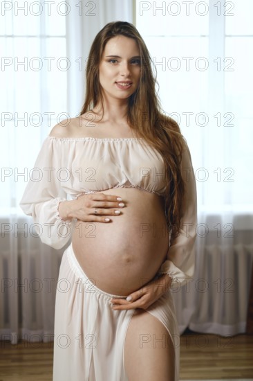 A pregnant woman stands gracefully in a softly lit room, wearing a flowing dress. She gently cradles her baby bump, showcasing her joy and anticipation of motherhood in a serene atmosphere