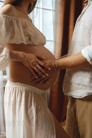 A pregnant woman in a soft dress enjoys a loving moment with her partner. They are inside a cozy room with natural light, highlighting the anticipation of their new arrival