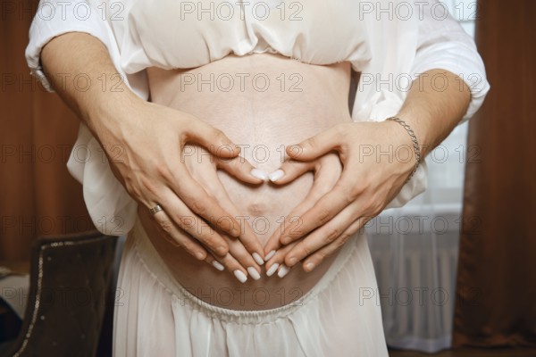 Two hands form a heart shape over a pregnant belly in a warm, softly lit room. The parents express joy and anticipation for their baby's arrival as they share a tender moment together