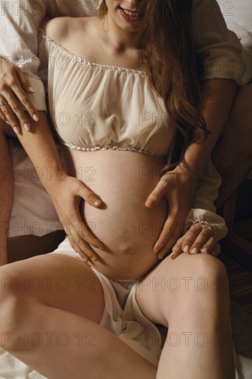 A couple sits on a soft bed, with one partner gently cradling the pregnant belly. The intimate atmosphere radiates love and joy, showcasing a special moment of connection before childbirth