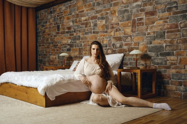 A pregnant woman sits elegantly in a warm-toned bedroom, showcasing her baby bump. The room features a wooden bed, soft lighting, and a serene atmosphere, ideal for relaxation