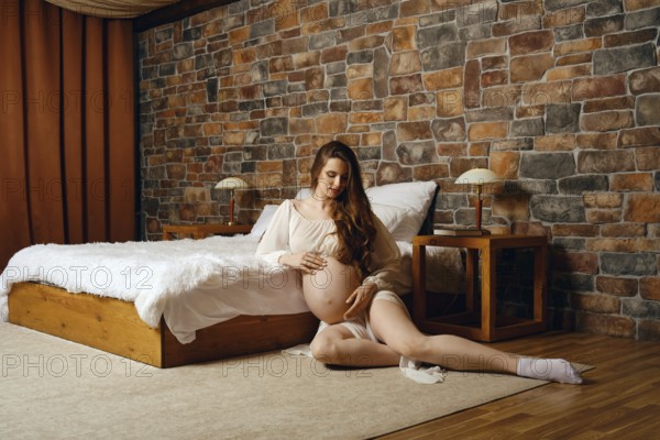 A pregnant woman sits in a warm, inviting bedroom, gently cradling her baby bump. The space features a wooden bed and rustic stone walls, creating a serene atmosphere for reflection