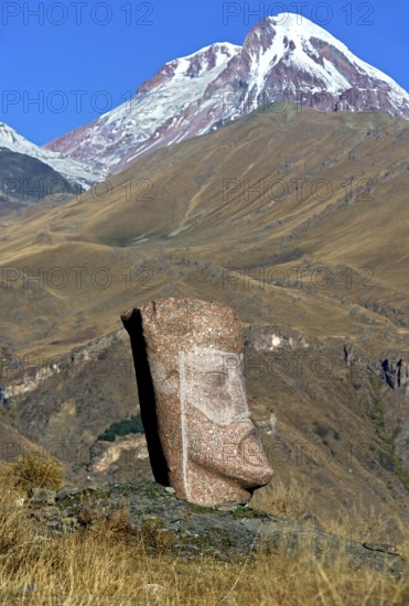 Stone sculpture of a male head, open-air exhibition Stone Heads of Sno by sculptor Merab Piranishvili, the snow-covered peak Kazbek behind, mountain village Sno, Kazbegi municipality in the Mtskheta-Mtianeti region, Georgia