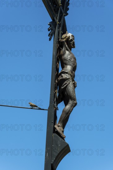 A detailed sculpture depicting Jesus on a cross rises against a bright blue sky. A small bird can be seen perched on a wire, adding life to the serene scene of religion and nature, Puy de Dome, Auvergne Rhone Alpes, France