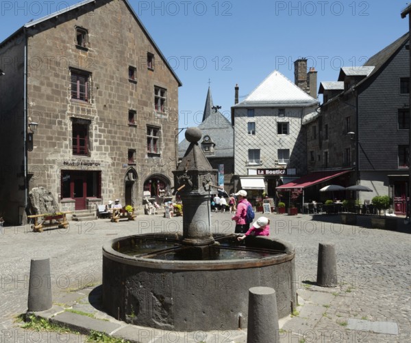 Visitors gather around a historic fountain in Besse en Chandesse, enjoying the warm sunshine and exploring the picturesque architecture of the village in Auvergne, France