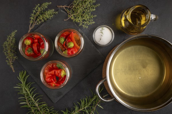 Glasses filled with chilli peppers next to a saucepan with vinegar, oil and herbs on a dark background