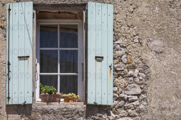 A window with blue shutters in a rustic house wall with flowering potted plants, Provence, France