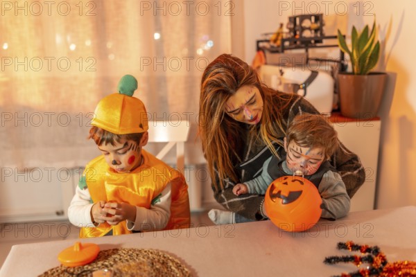 Mother and children wearing halloween costumes are sitting at a table at home, playing with pumpkin decorations and candies