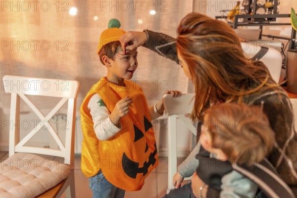 Mother applying halloween makeup on her son wearing a pumpkin costume, while holding her baby