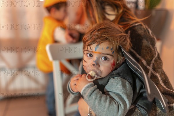 Toddler wearing a halloween costume with painted face and pacifier, held by mother, with another child in the background