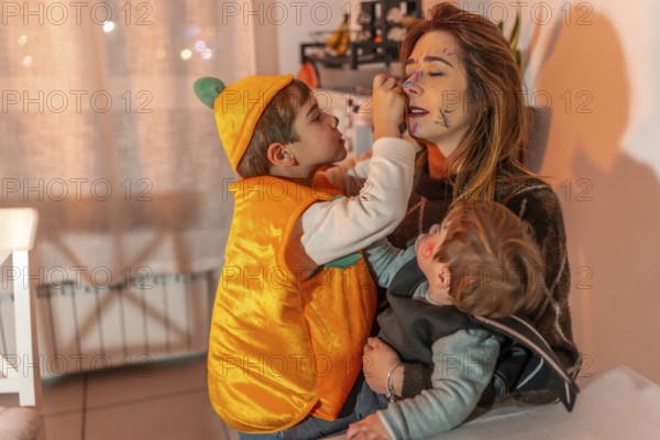 Two young brothers in halloween costumes playfully applying makeup on their mother's face in a festive living room