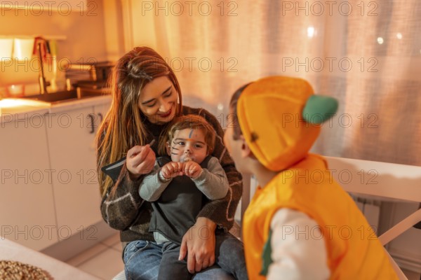 Mother applying halloween makeup on her toddler son dressed up as a cat while his brother dressed up as a pumpkin watches
