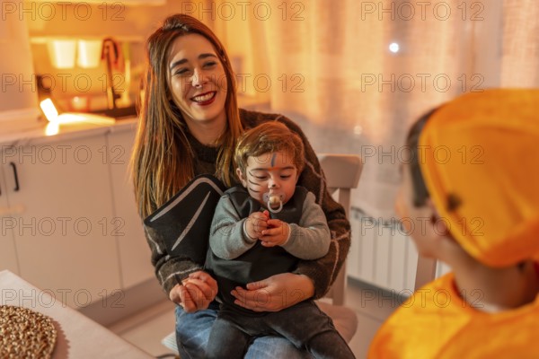 Mother holding her baby dressed up as a bat while smiling at her other child dressed in orange costume, celebrating halloween