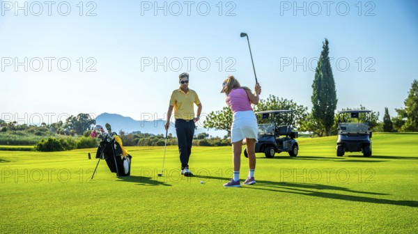 Golf instructor coaching female golfer on driving range, with golf carts in background on sunny day