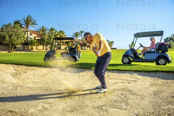 Golfer hitting ball out of sand trap with golf carts and other golfers in background on sunny day