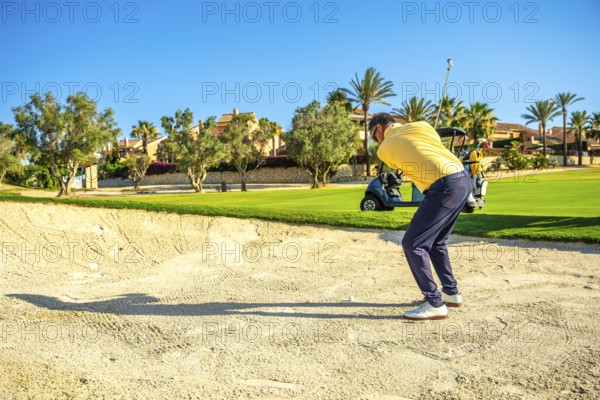 Golfer concentrating on hitting ball out of sand trap on a beautiful sunny day, with golf cart in background