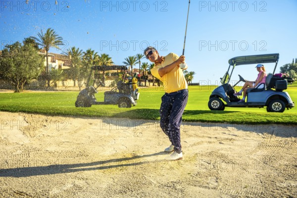 Male golfer hitting ball out of sand trap with golf cart and other players in background on sunny day