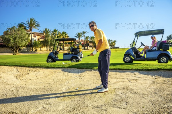 Golfer focuses on a bunker shot while others relax in golf carts on a bright, sunny day at a lush golf course with palm trees