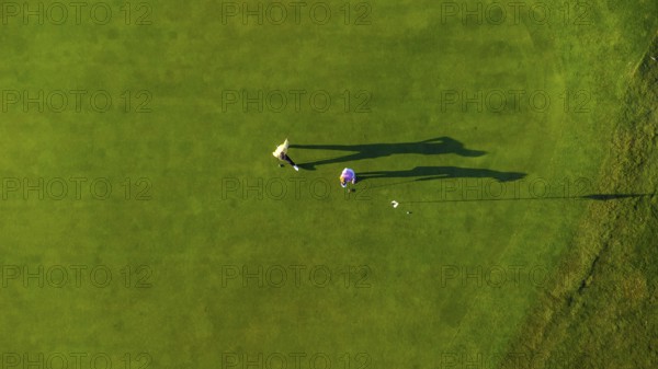 Two golfers are putting on a vibrant green golf course, seen from an aerial perspective, creating long shadows in the afternoon sun