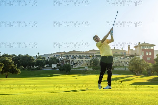 Professional golfer swinging his club, hitting a golf ball on a lush green course, with a luxury hotel visible in the background during a sunny day