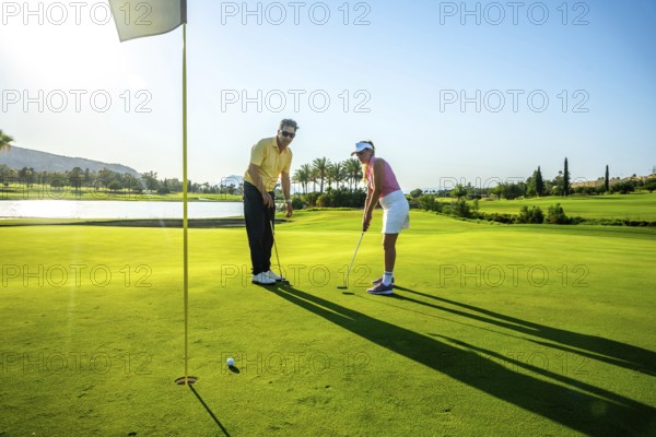 Two golfers practicing putting on a bright, sunny day at a lush golf course, surrounded by palm trees and a serene water feature