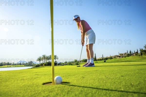 Woman focusing on a putt on a bright, sunny day at a lush golf course, with a ball near the hole and scenic landscape in the background