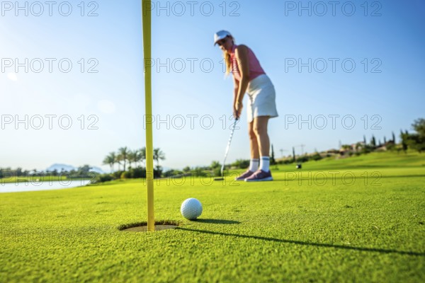 Woman golfer putting a ball on the green, aiming for the hole under a bright sun at a picturesque golf course on a summer day
