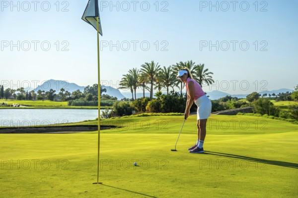 Woman golfer concentrating on putting on the green of a beautiful golf course with palm trees, a lake, and mountains