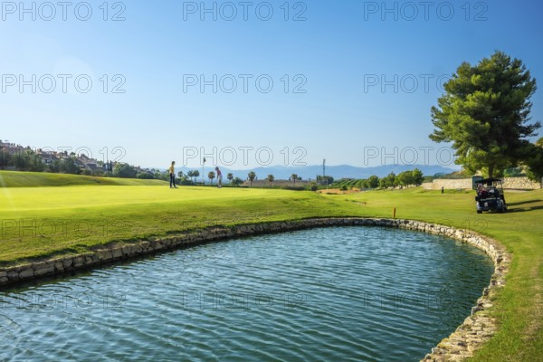 Two golfers are playing on a sunny golf course with a water hazard and golf cart in the background
