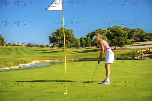 Female golfer concentrating on putting the ball on a sunlit day, enjoying the beauty of a lush golf course surrounded by nature