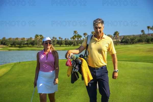 Couple enjoying a sunny day playing golf, walking on a beautiful green golf course, carrying golf clubs and enjoying the sport
