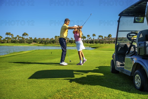 Golf instructor guiding female golfer on a sunny course, improving her swing technique near a golf cart and a water hazard