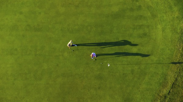 Two golfers are playing on a perfectly manicured green golf course on a sunny day, casting long shadows on the grass, seen from a high angle aerial view
