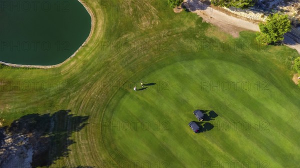 Scenic aerial view of a golf course featuring two players walking near golf carts on a lush green fairway beside a pond under clear skies
