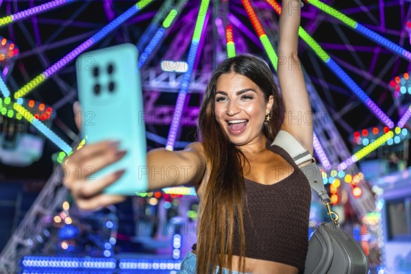 Young woman taking a selfie with smartphone in front of a colorful ferris wheel at an amusement park, enjoying the vibrant atmosphere