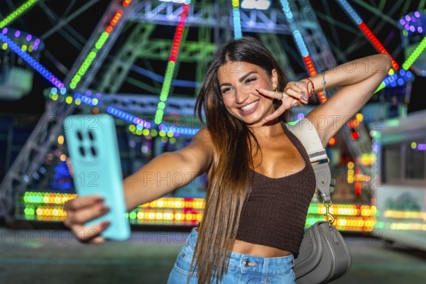 Happy young woman taking a selfie and making peace sign at amusement park with colorful ferris wheel in background at night