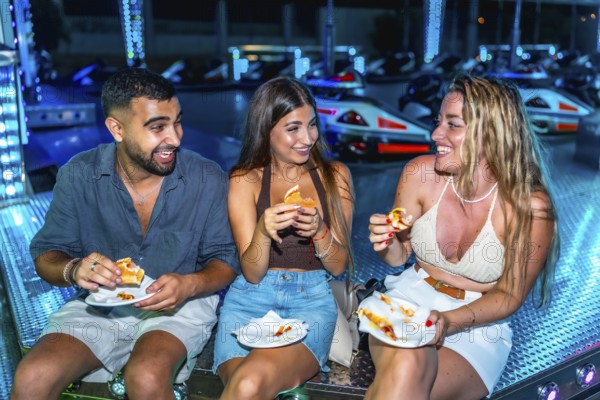 Three friends are enjoying sandwiches while sitting on a bumper cars platform, illuminated by colorful lights at night