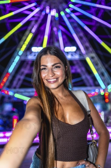 Happy young woman taking a selfie with a colorful ferris wheel in the background at an amusement park