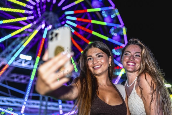 Two happy women taking a selfie with smartphone in front of illuminated ferris wheel at an amusement park