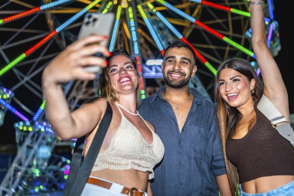 Three happy friends taking a selfie with smartphone in front of a colorful ferris wheel at an amusement park at night