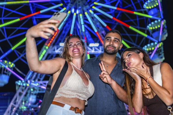 Three friends taking a selfie with a smartphone in front of a brightly lit ferris wheel at an amusement park at night