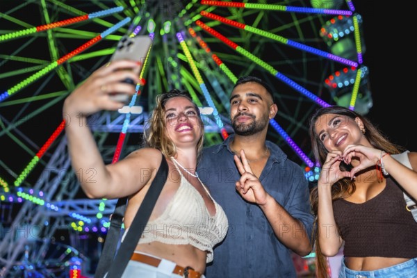 Three friends taking a selfie with smartphone in front of a colorful ferris wheel, enjoying nightlife and summer holidays