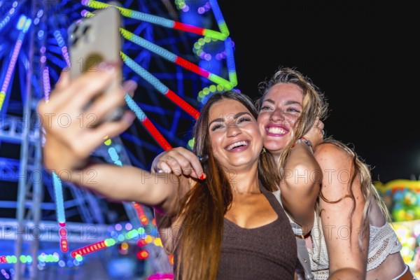 Two cheerful young women taking a selfie in front of a brightly lit ferris wheel at an amusement park at night, enjoying their summer vacation