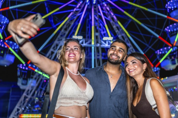 Three happy friends taking a selfie with smartphone in front of a colorful ferris wheel at an amusement park