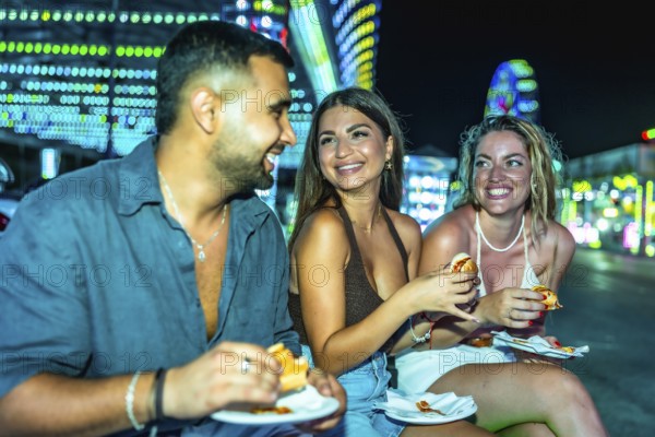Happy friends sharing delicious hot dogs while enjoying the vibrant atmosphere of an amusement park at night, filled with laughter and joy