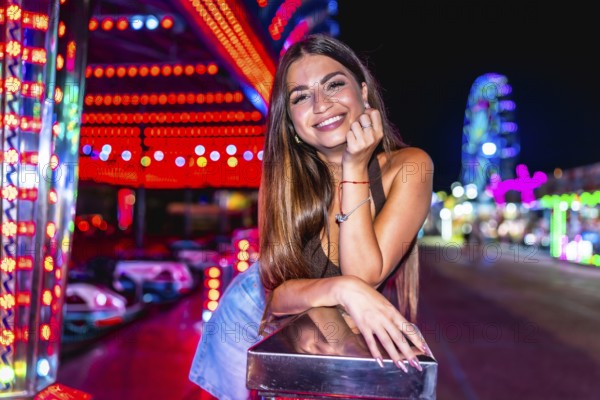 Happy woman enjoying night at amusement park, leaning on railing with colorful bumper cars and ferris wheel in background