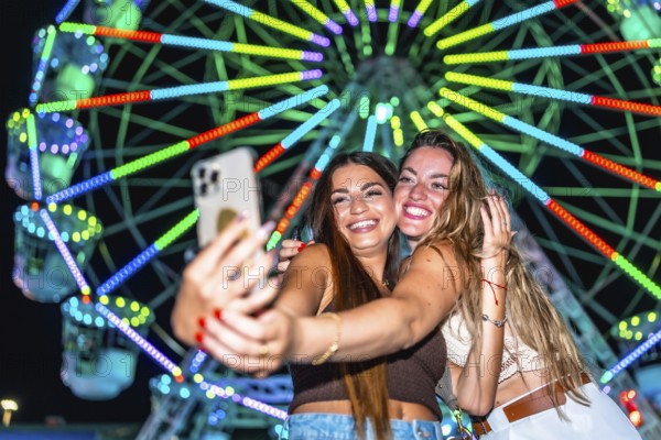 Two young women taking a selfie with a smartphone in front of a brightly lit ferris wheel at an amusement park at night, enjoying their time together