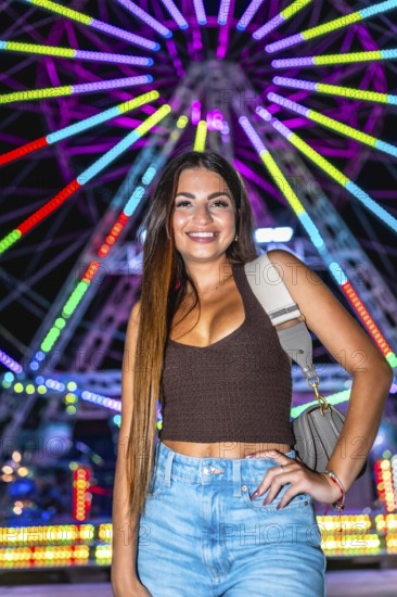 Happy young woman posing at night with illuminated ferris wheel in background, enjoying funfair attractions
