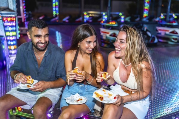 Three friends are sitting and laughing while eating hot dogs at an amusement park's bumper cars attraction